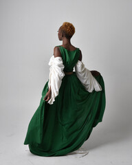 Full length portrait of pretty African woman wearing long green medieval fantasy gown, standing pose facing backwards on a light grey studio background.