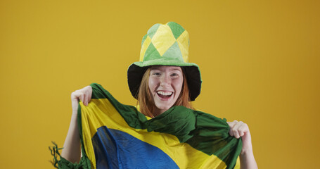 Brazil supporter. Brazilian redhead woman fan celebrating on soccer, football match on yellow background. Brazil colors. Wearing a t-shirt, flag and fan hat.