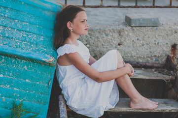 A charming teenager girl with long hair in a white dress sits leaning against a turquoise wooden boat. Mood spleen