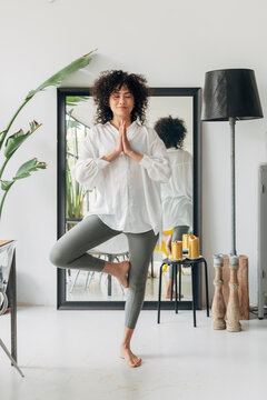 Young mixed race female with curly hair practicing yoga at home. Tree pose, hands in prayer.