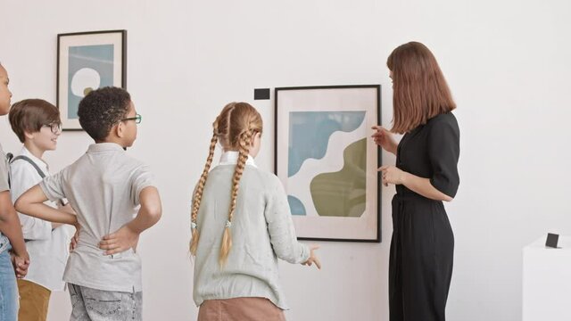 Medium long side view of multiethnic school students looking at paintings, blond-haired girl asking questions, young Caucasian woman wearing black outfit talking to children in art gallery