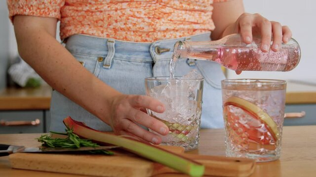 drinks and people concept - young woman making rhubarb cocktail and decorating it with sprig of rosemary at home kitchen
