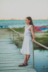 A cute teenage girl with long hair in a white dress stands and dreams on the sea promenade