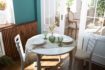 Green glasses and white dishes on the kitchen table, with green plant on living room interior apartment in the background