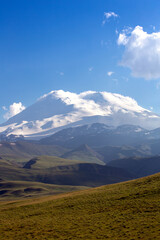 Fototapeta premium Snow-capped mountain peaks on a summer day. Elbrus on the background of green hills. Mountain landscape. The Caucasus Mountains. Blue sky with clouds. Russia.