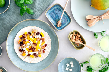 Flat lay of a delicious tortilla wrap with feta cheese, yellow paprika, raisins and cashew nuts with honey, surrounded by blue-green ceramic plates and bowls with a mango in the right hand corner.