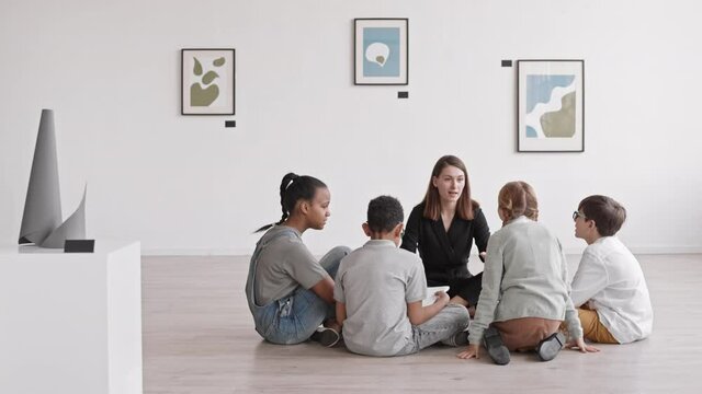 Long shot of young female Caucasian teacher and diverse student sitting in circle on floor of art gallery, having lesson