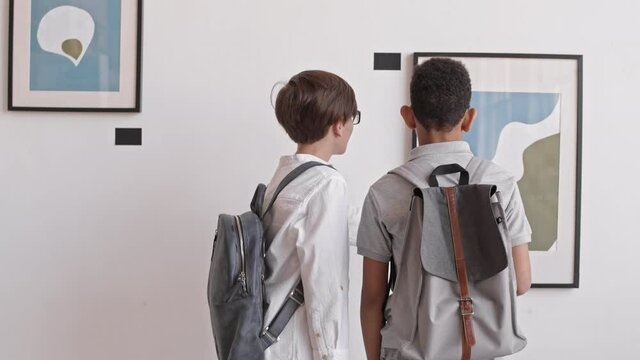 Rear view of two multiethnic school-aged boys wearing backpacks, observing paintings in art gallery