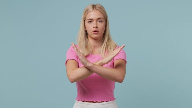 Serious strict severe frowning sad young blonde woman 20s wears pink t-shirt say no hold palm folded crossed hands in stop gesture isolated on pastel plain light blue color background studio portrait
