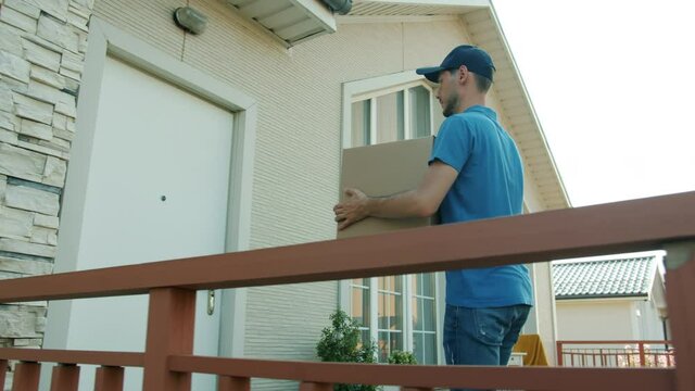 Delivery guy in uniform is carrying carcel to house and knocking on door waiting for client outside working alone. Modern commerce and e-shopping concept.