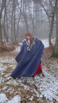 Funny woman with red dreadlocks in viking costume with hatchet and mantle runner into snow forest. Amateur shooting of cosplayer's retreating person from back. Winter atmosphere in wood.
