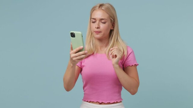 Smiling young blonde woman 20s wears pink t-shirt doing selfie shot on mobile phone post photo on social network blow send air kiss isolated on pastel plain light blue color background studio portrait