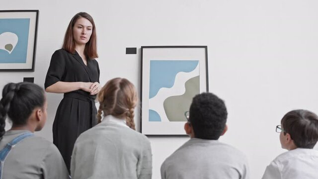 Low angle rear view of four school students sitting on floor of art gallery, young Caucasian woman presenting them painting, answering questions