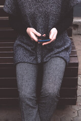 Young woman in a cozy gray-blue vest spends time outdoors in the city. Female sits on a bench with mobile phone. Lifestyle portrait of girl