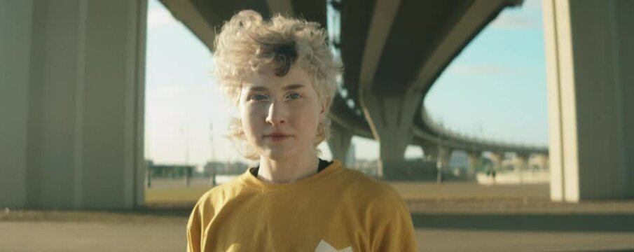 Zoom in anamorphic portrait shot of young beautiful female dancer in streetwear standing outdoors under urban bridge and looking at camera