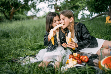 Joyful little girls in summer outfit sitting on checkered blanket and biting one banana from ends. Happy pretty sisters spending free time outdoors with fun.