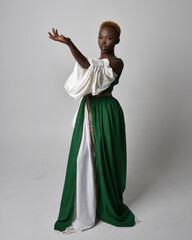 Full length portrait of pretty African woman wearing long green medieval fantasy gown, standing and dancing pose on a light grey studio background.