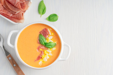 Top view of salmorejo spanish soup made of pureed tomatoes and bread decorated with basil leaf and chopped boiled egg served in bowl with jamon or ham and spoon on wooden table. Image with copy space