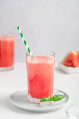 Red refreshing cold watermelon smoothie drink served in drinking glass with ice cubes and paper straw on round plate with ripe fruit slices and basil leaf on white wooden table ib hot summer season