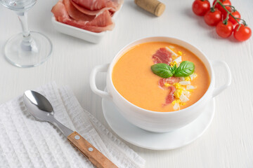 Salmorejo spanish soup made of pureed tomatoes and bread decorated with basil leaf and chopped boiled egg served in bowl with jamon or ham, spoon and glass of wine on white wooden table in summer