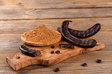 Dry carob pods and carob powder over wooden background. Organic healthy ingredient for vegan vegetarian food and drinks, close up - obrazy, fototapety, plakaty