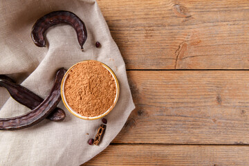 Dry carob pods and carob powder on linen napkin over wooden background. Organic healthy ingredient for vegan vegetarian food and drinks, copy space