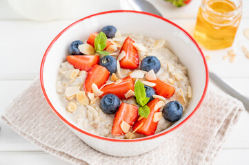 Bowl of oatmeal porridge with blueberries, strawberries, almond petals and honey on a white wooden background. Healthy food for breakfast, top view, copy space.