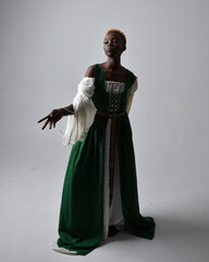 Fototapeta premium Full length portrait of pretty African woman wearing long green medieval fantasy gown, standing and dancing pose on a light grey studio background.