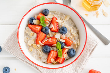 Bowl of oatmeal porridge with blueberries, strawberries, almond petals and honey on a white wooden background. Healthy food for breakfast, top view, copy space.