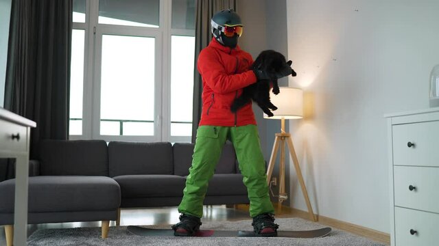 Funny video. Man dressed as a snowboarder depicts snowboarding on a carpet in a cozy room. He plays with a black fluffy cat. Waiting for a snowy winter