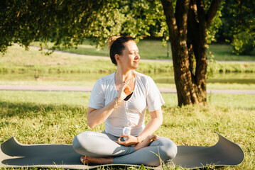 Happy young woman practicing yoga and meditation sitting in lotus position on yoga mat in park at sunrise. Portrait of smiling professional yoga woman in morning outdoors. Healthy lifestyle concept