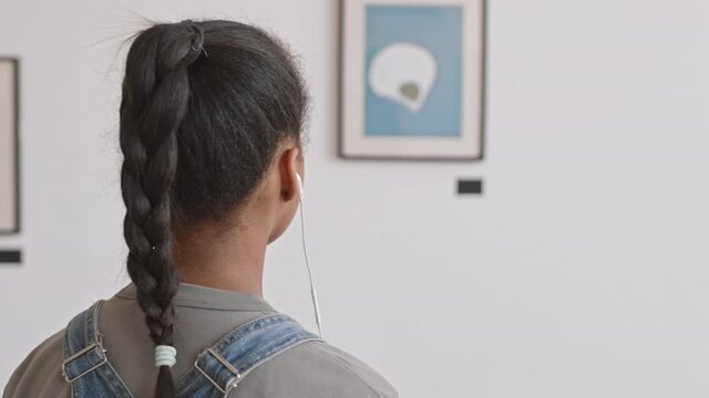 Rear view of African school-aged girl wearing wired headphones, listening to audio guide while looking at contemporary paintings on exhibition