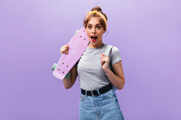 Surprised woman in stylish outfit holds longboard. Lovely girl with yellow bandana in denim skirt with black wide belt posing.