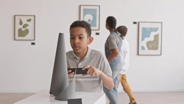 Medium close-up of twelve-year-old African boy taking pictures of art object on exhibition in museum, using smartphone