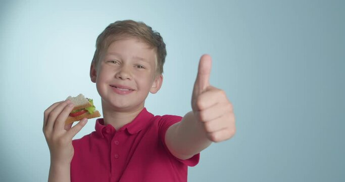 Little boy eating tasty sandwich and showing thumb-up on color background