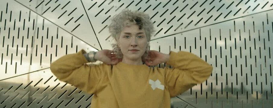 Zoom in anamorphic shot of young female dancer in streetwear standing in front of perforated wall, looking at camera and dancing vogue