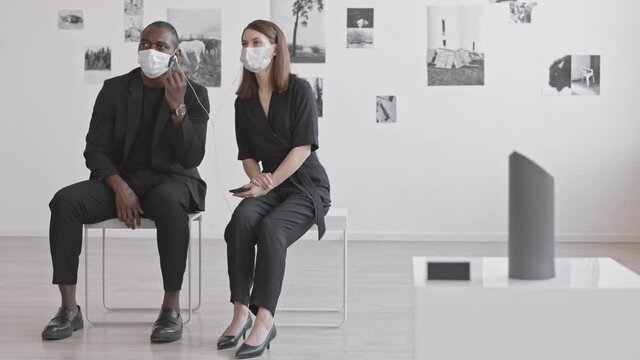 Full shot of young African man and Caucasian woman wearing black clothes and medical masks, sitting in contemporary art gallery, listening to audio guide in wired earphones