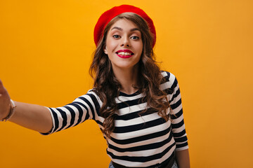 Smiling lady in bright beret makes selfie on orange background. Charming woman with curly hair with red lips posing on isolated backdrop..