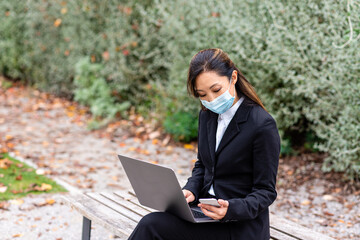 telecommunting in a pubblic park, adult japanese business woman typing on her laptop, successful and confident female manager wearing protective mask and use smartphone, the new normal lifestyle