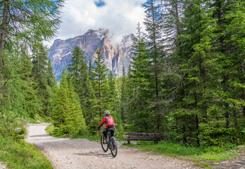 nice and active senior woman riding her electric mountain bike on the Pralongia Plateau in the Alta Badia Dolomites with awesome Sasso die Santa Cruce summit in Backg, South Tirol and Trentino, Italy