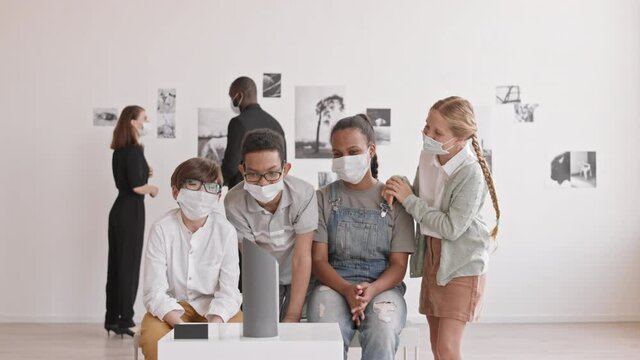 Medium long of African and Caucasian boys and girls wearing medical masks carefully looking at modern art exhibit in contemporary gallery, man and woman joining them