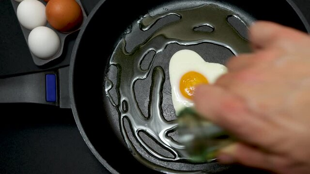A frying pan with a fried egg in the shape of a heart and next to it half a dozen white and brown eggs, all on a black background, healthy cooking and cholesterol concept