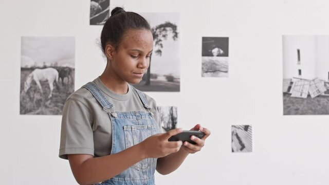 Waist-up of teenage African girl standing in museum at contemporary photography exhibition, talking selfie with smartphone, turning around