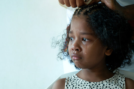 Close up African hairstylist braided hair of afro little girl, young girl makes a painful expression as she gets braided African-style hair