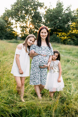 Beautiful pregnant woman with two cute daughters spending free time on nature. Little sisters standing near mother and embracing pregnant tummy. Love and family concept.