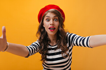 Surprised lady in red beret makes selfie on orange background.Modern woman with curly hair with bright lips in striped clothes posing..