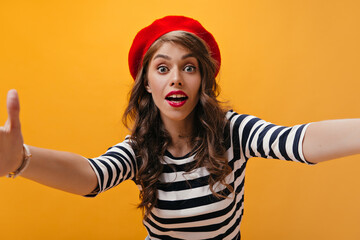 Naklejka premium Surprised lady in red beret makes selfie on orange background.Modern woman with curly hair with bright lips in striped clothes posing..