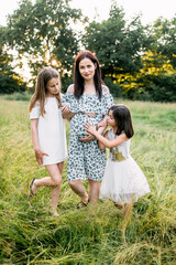 Fototapeta premium Beautiful pregnant woman with two cute daughters spending free time on nature. Little sisters standing near mother and embracing pregnant tummy. Love and family concept.