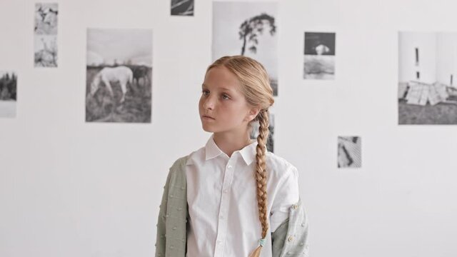 Waist-up of blond-haired Caucasian girl wearing two braids standing in museum at contemporary photography exhibition, talking pictures with smartphone