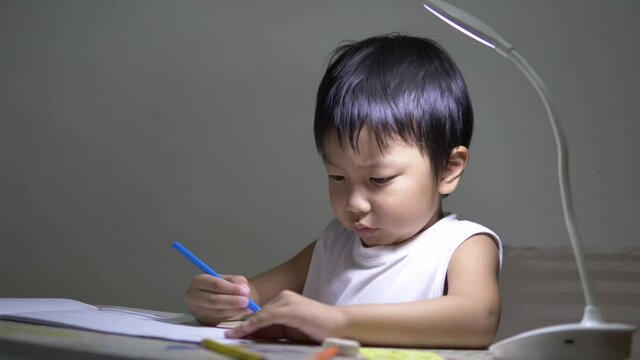 Asian cute child boy writing on paper by pencil. Kid studying and doing homework with concentrate face in room with lighting lamp background. Concept of children education, school at home.